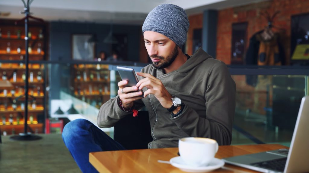 Young handsome hipster man with beard sitting in cafe talking mobile phone, holding cup of coffee